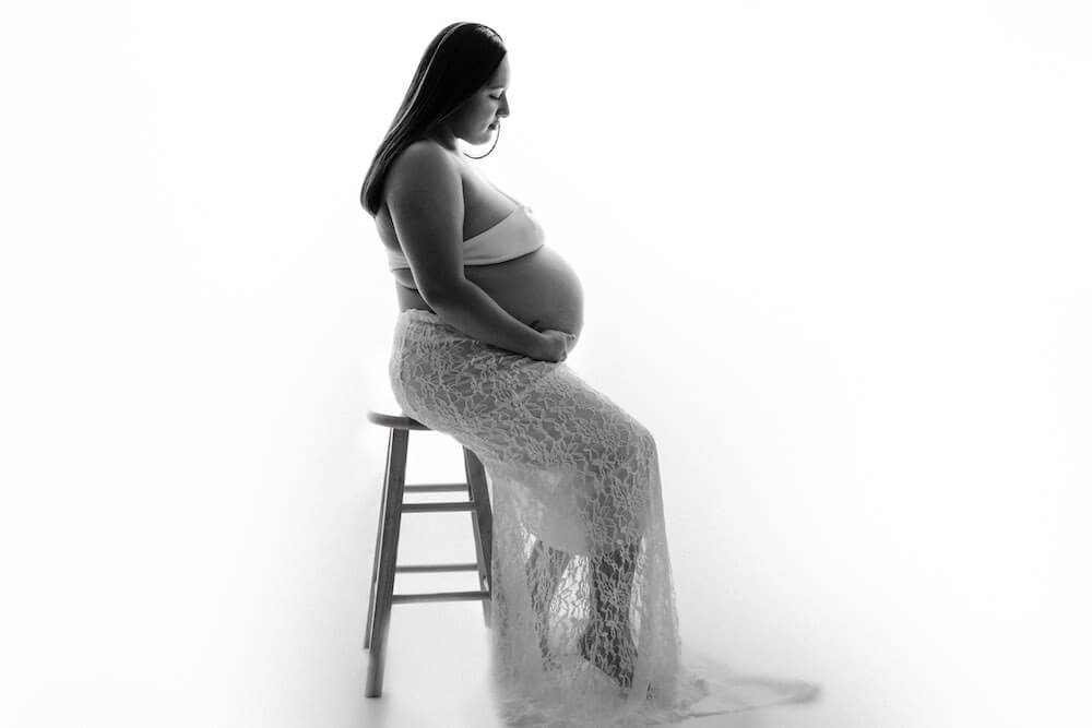 A black and white maternity photograph of a woman seating on a stool